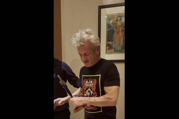 Ein Mann mit zerzausten Haaren hält eine Trophäe und trägt ein schwarzes T-Shirt mit auffälligem Aufdruck.