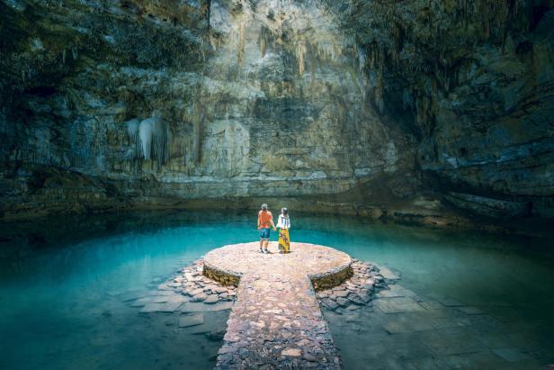 Zwei Menschen stehen auf einer Plattform in einer Cenote mit türkisblauem Wasser und Felswänden.
