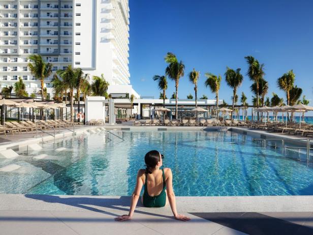Eine Frau sitzt in einem grünen Badeanzug an einem Pool. Im Hintergrund sind Palmen und ein Hotel mit mehreren Etagen zu sehen. 