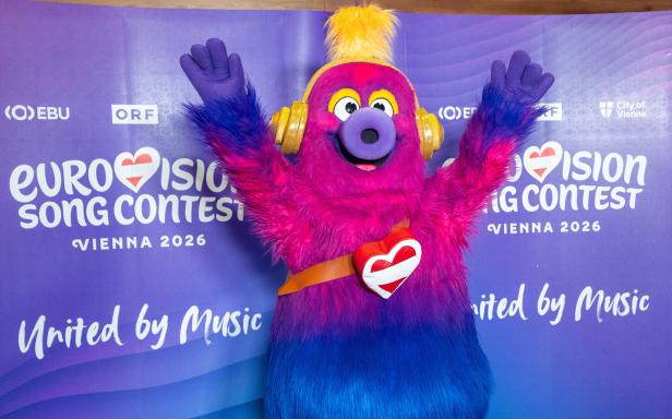 Ein farbenfrohes Maskottchen mit Kopfhörern posiert vor einem Eurovision Song Contest Vienna 2026 Banner.