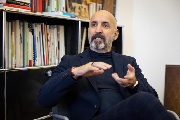 Ein Mann mit Bart und Glatze sitzt vor einem Bücherregal und gestikuliert mit den Händen, als ob er etwas erklärt.