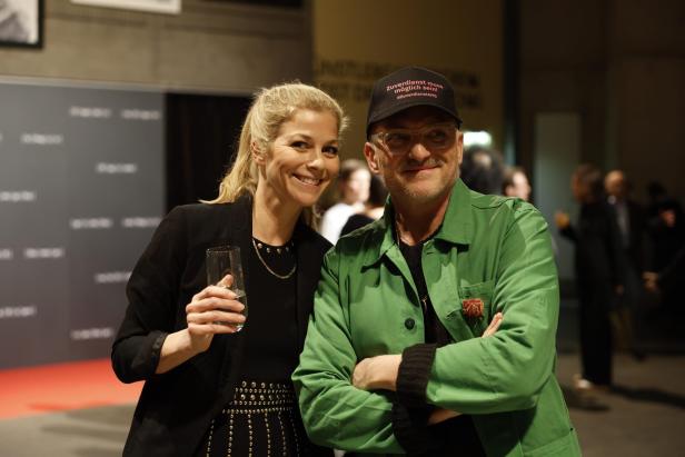 Eine Frau mit blondem Pferdeschwanz hält ein Glas und steht lächelnd neben einem Mann mit Brille und grüner Jacke.