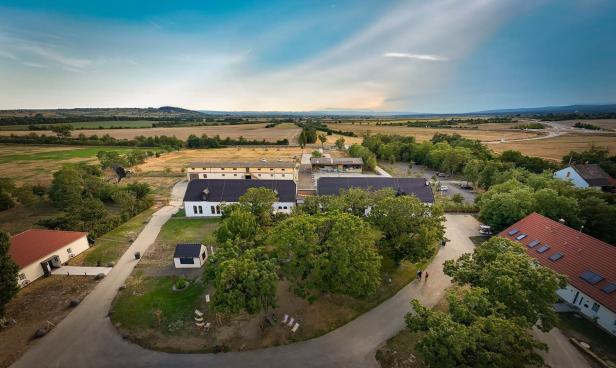Mehrere Gebäude mit roten und dunklen Dächern sind von Bäumen umgeben, dahinter weite Felder und ein weiter Horizont.