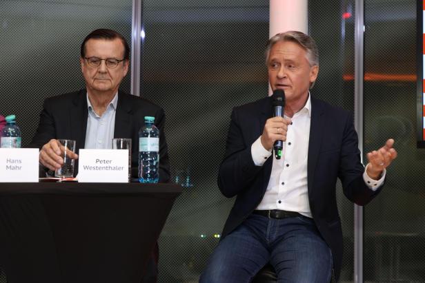 Zwei Männer sitzen bei einer Podiumsdiskussion, einer spricht ins Mikrofon.