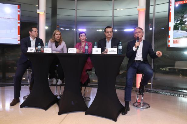 Fünf Personen sitzen bei einer Podiumsdiskussion an Tischen mit Namensschildern.