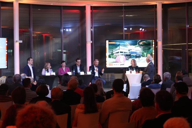 Podiumsdiskussion mit mehreren Teilnehmenden vor Publikum und großem Bildschirm im Hintergrund.