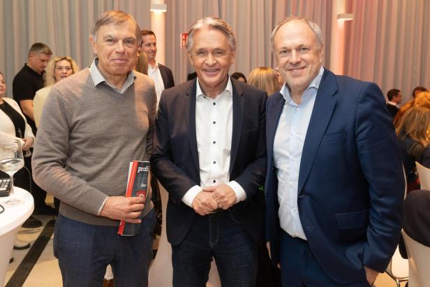 Drei Männer in Anzügen stehen lächelnd bei einer Veranstaltung im Gespräch zusammen.