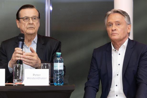 Zwei Männer im Anzug sitzen an einem Tisch, einer spricht in ein Mikrofon.