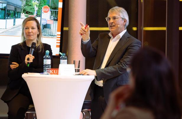 Zwei Personen diskutieren auf einer Bühne an einem Stehtisch mit Mikrofon und Wasserflaschen.