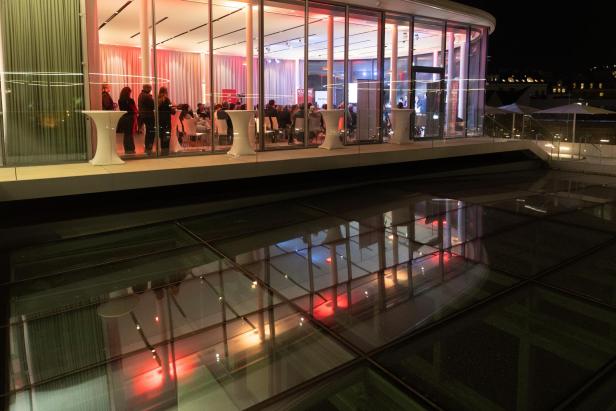 Menschen sitzen und stehen bei einer abendlichen Veranstaltung in einem verglasten Saal.