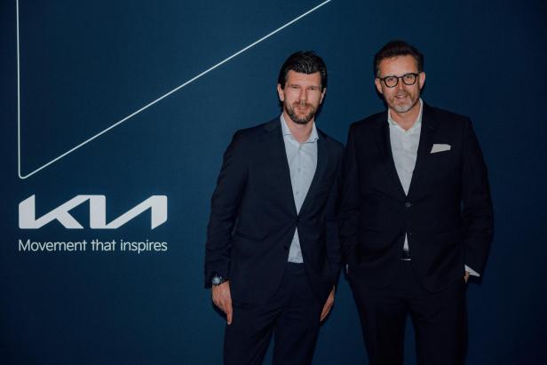 Zwei Männer in Anzügen posieren vor einer blauen Wand mit Kia-Logo und Slogan.