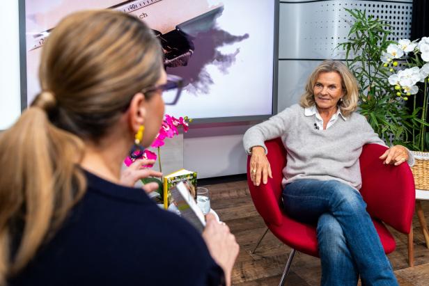 Zwei Frauen unterhalten sich in einem modernen Raum, eine sitzt entspannt in einem roten Sessel, umgeben von Pflanzen.