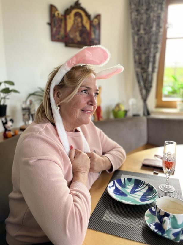 Eine lächelnde Frau mit Hasenohren sitzt an einem Tisch mit Geschirr und einem Sektglas.