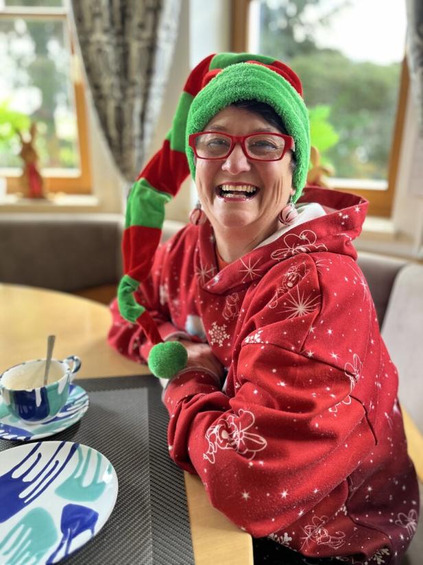 Eine lachende Frau mit Brille und Wichtelmütze sitzt an einem Tisch mit Geschirr.