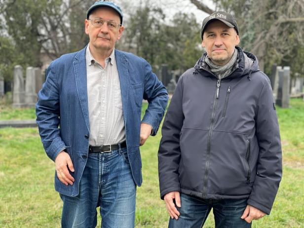 Zwei Männer stehen nebeneinander auf einer Wiese mit Grabsteinen im Hintergrund.