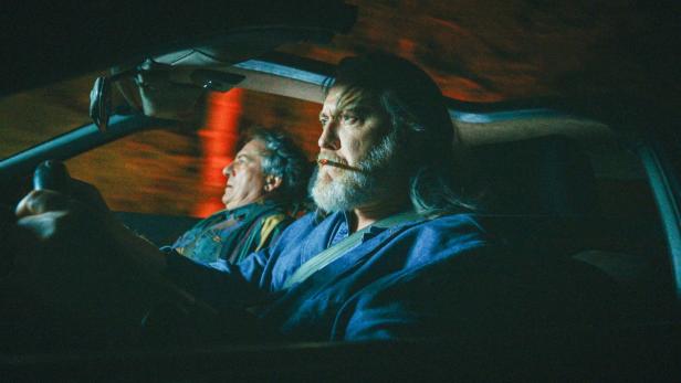 Zwei Männer sitzen nachts im Auto, der Fahrer mit ernstem Blick und Zigarre im Mund, der Beifahrer wirkt angespannt.