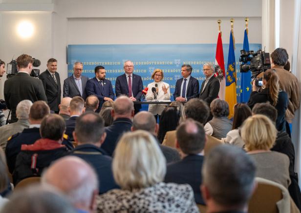 Mehrere Personen stehen an einem Rednerpult vor Publikum, im Hintergrund sind Fahnen und ein blaues Banner mit Aufschrift.