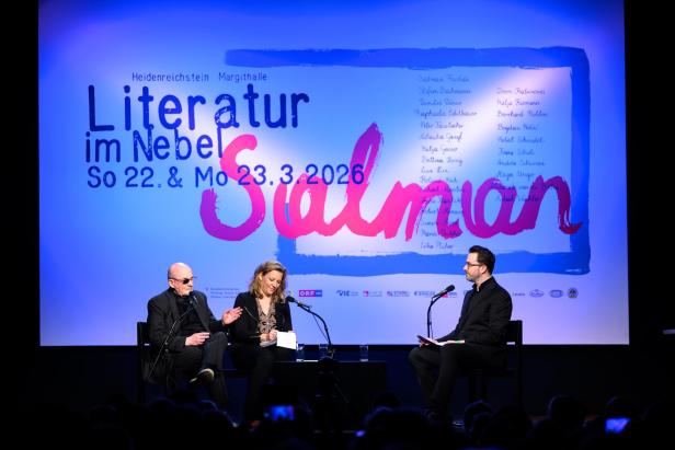 Drei Personen sitzen auf einer Bühne und diskutieren vor einer großen, bunt beleuchteten Wand mit Veranstaltungsankündigung.