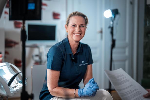 Zahnärztin in Praxisumgebung mit blauer Uniform und Handschuhen lächelt in die Kamera.