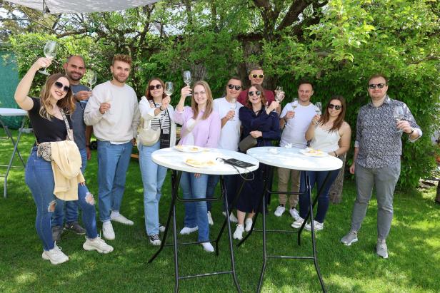 Elf Personen stehen im Garten, halten leere Weingläser hoch und lächeln gemeinsam in die Kamera.