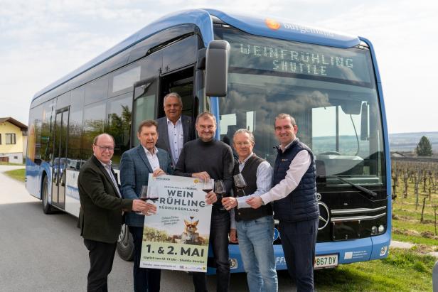 Sechs Männer stehen vor einem blauen Bus mit der Aufschrift „Weinfrühling Shuttle“ und halten Weingläser sowie ein Veranstaltungsplakat.