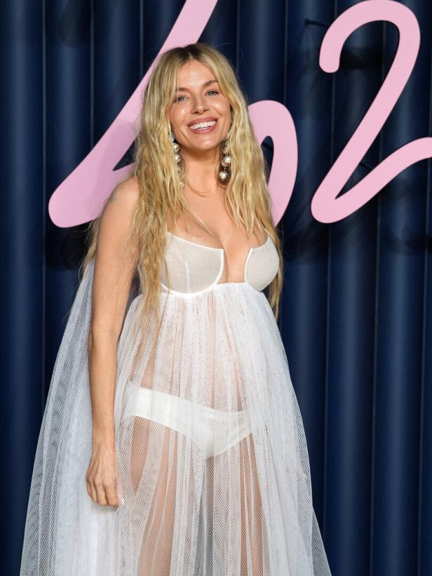 Sienna Miller lächelt in einem weißen, transparenten Kleid vor einem blauen Hintergrund.