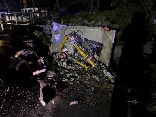 Ein zerstörtes gelbes Metallobjekt liegt vor einer beschädigten Betonwand, umgeben von Trümmern und Autoteilen.