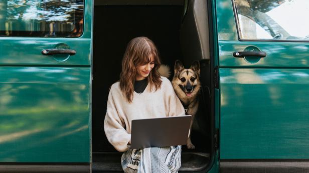 Eine junge Frau sitzt mit einem Laptop und einem Hund in der geöffneten Tür eines grünen Transporters und arbeitet flexibel ortsunabhängig.