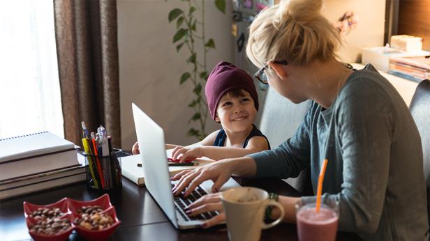 Eine Frau arbeitet am Laptop zu Hause, während ein Kind neben ihr sitzt und sie anlächelt, was die Vereinbarkeit von Studium, Beruf und Familie verdeutlicht.