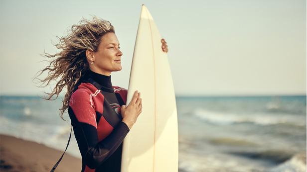 Eine Frau im Neoprenanzug steht mit einem Surfbrett am Strand und blickt zufrieden aufs Meer, was für Flexibilität und Selbstbestimmung im Alltag steht.