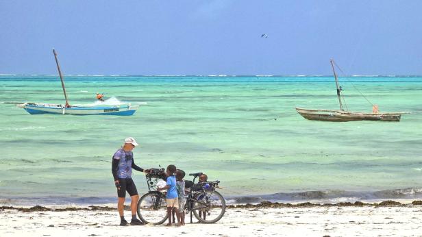 Mann mit Fahrrad und drei Kindern am weißen Sandstrand vor türkisblauem Meer und Segelbooten.