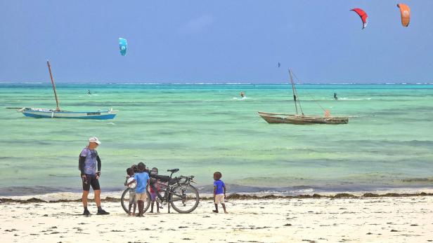 Mann mit Fahrrad und drei Kindern am weißen Sandstrand vor türkisblauem Meer und Segelbooten.