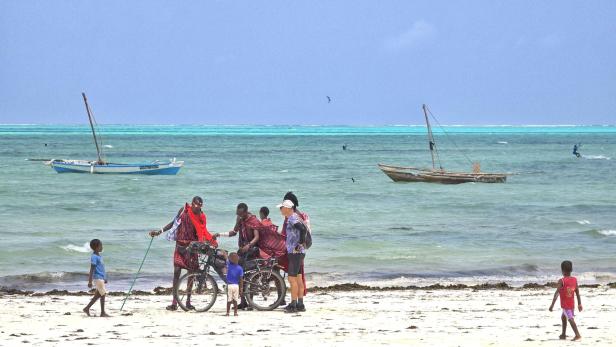 Mann mit Fahrrad und drei Kindern am weißen Sandstrand vor türkisblauem Meer und Segelbooten.