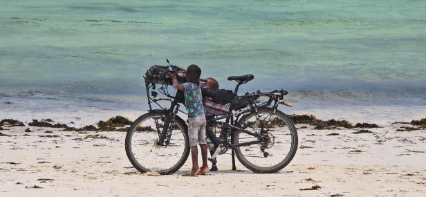 Mann mit Fahrrad und drei Kindern am weißen Sandstrand vor türkisblauem Meer und Segelbooten.