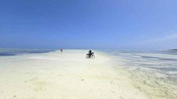 Mann mit Fahrrad und drei Kindern am weißen Sandstrand vor türkisblauem Meer und Segelbooten.
