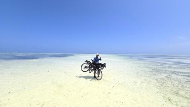 Mann mit Fahrrad und drei Kindern am weißen Sandstrand vor türkisblauem Meer und Segelbooten.