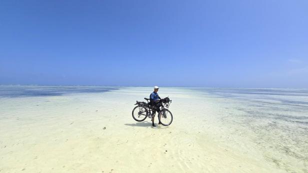 Mann mit Fahrrad und drei Kindern am weißen Sandstrand vor türkisblauem Meer und Segelbooten.