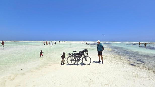 Mann mit Fahrrad und drei Kindern am weißen Sandstrand vor türkisblauem Meer und Segelbooten.