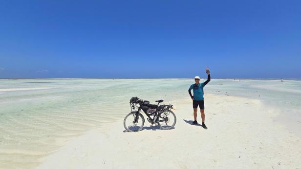Mann mit Fahrrad und drei Kindern am weißen Sandstrand vor türkisblauem Meer und Segelbooten.