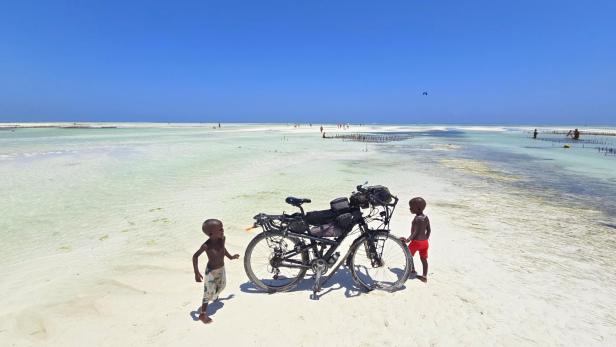 Mann mit Fahrrad und drei Kindern am weißen Sandstrand vor türkisblauem Meer und Segelbooten.