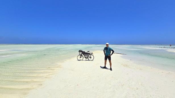 Mann mit Fahrrad und drei Kindern am weißen Sandstrand vor türkisblauem Meer und Segelbooten.
