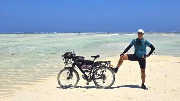 Mann mit Fahrrad und drei Kindern am weißen Sandstrand vor türkisblauem Meer und Segelbooten.