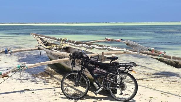 Mann mit Fahrrad und drei Kindern am weißen Sandstrand vor türkisblauem Meer und Segelbooten.