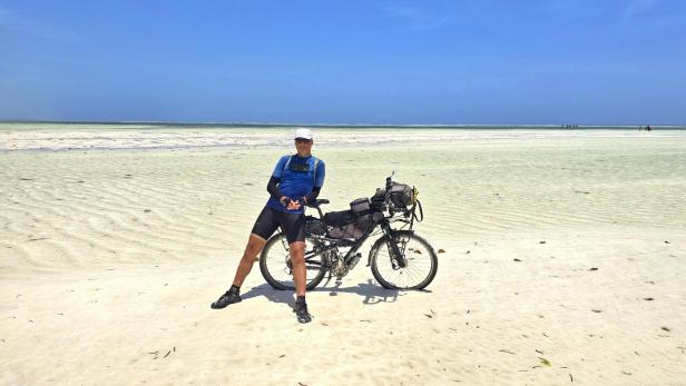 Mann mit Fahrrad und drei Kindern am weißen Sandstrand vor türkisblauem Meer und Segelbooten.