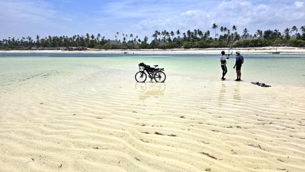 Mann mit Fahrrad und drei Kindern am weißen Sandstrand vor türkisblauem Meer und Segelbooten.