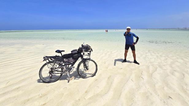 Mann mit Fahrrad und drei Kindern am weißen Sandstrand vor türkisblauem Meer und Segelbooten.