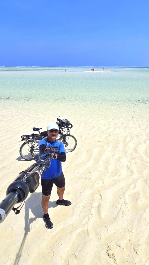 Mann mit Fahrrad und drei Kindern am weißen Sandstrand vor türkisblauem Meer und Segelbooten.