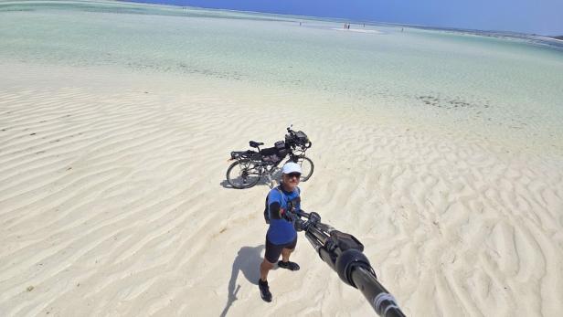 Mann mit Fahrrad und drei Kindern am weißen Sandstrand vor türkisblauem Meer und Segelbooten.