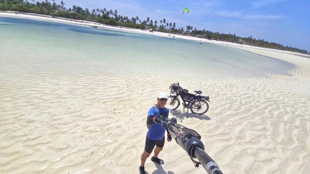 Mann mit Fahrrad und drei Kindern am weißen Sandstrand vor türkisblauem Meer und Segelbooten.