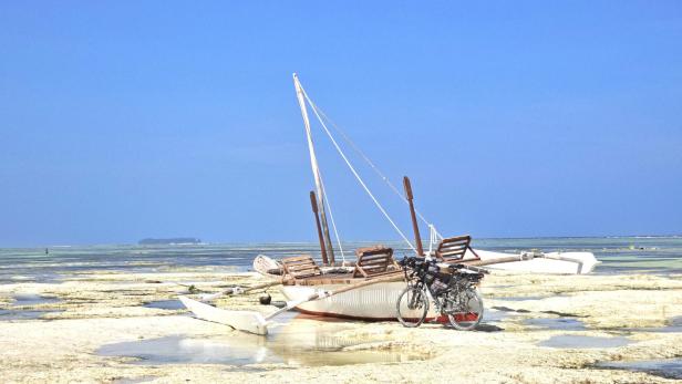 Mann mit Fahrrad und drei Kindern am weißen Sandstrand vor türkisblauem Meer und Segelbooten.